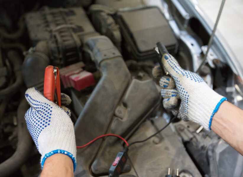 car service, repair, maintenance and people concept - auto mechanic man with cleats charging battery at workshop